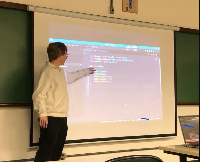Me at the projector in a PUCPR classroom, pointing at a FastAPI file on screen while walking the room through it.