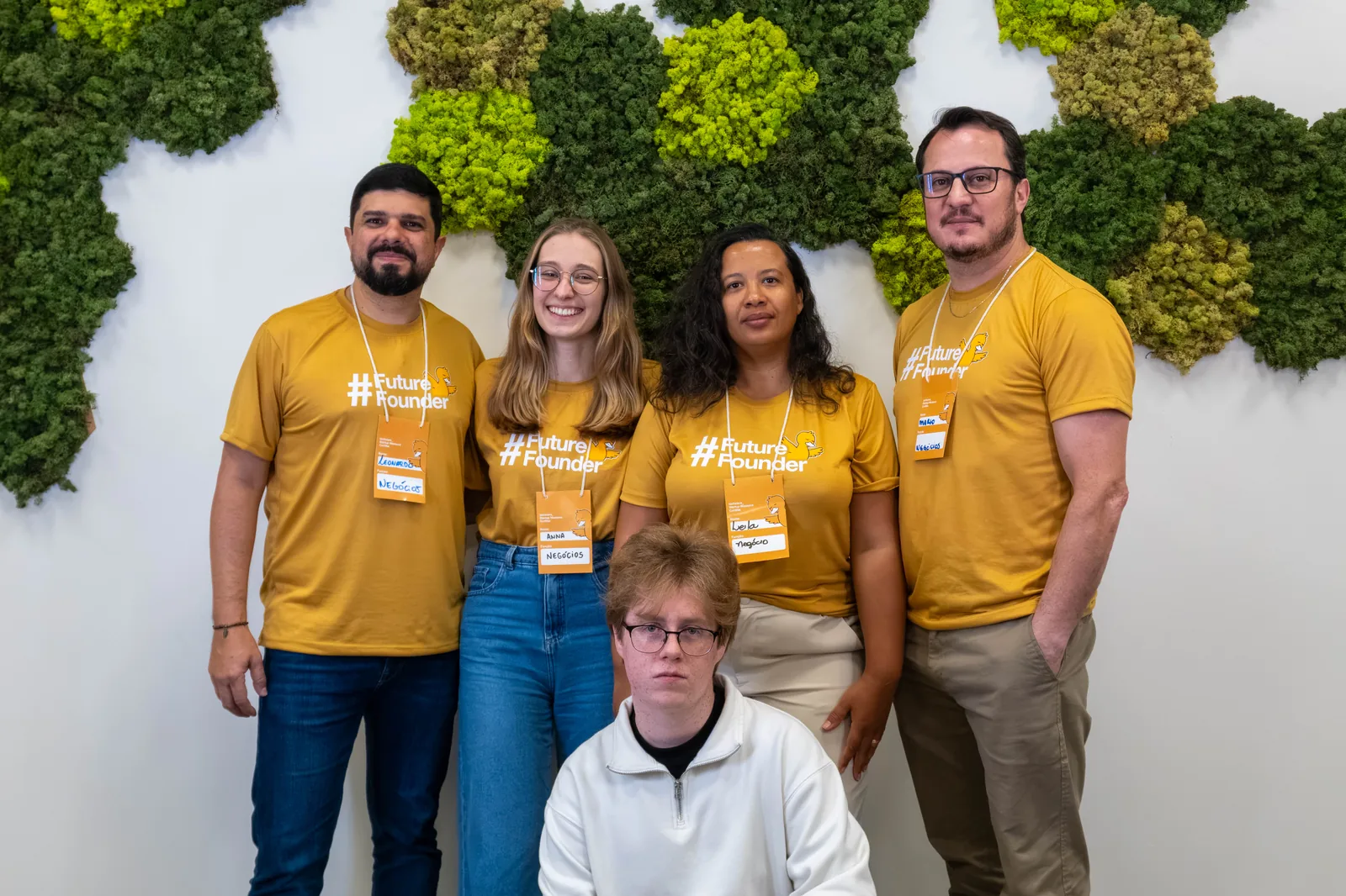 Five FuelCheck teammates posing in Startup Weekend shirts in front of a moss wall.